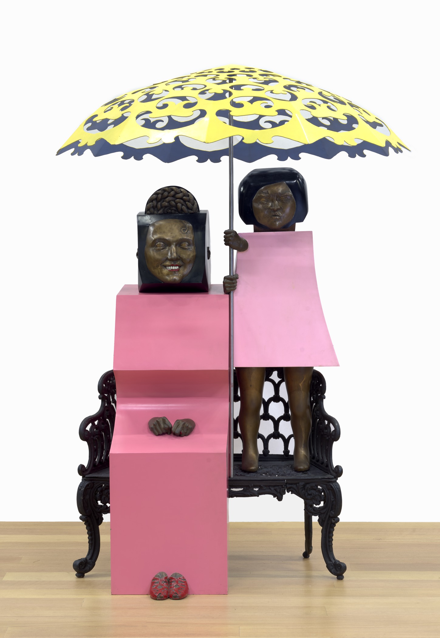 A Pop Art sculpture of a woman and young girl sitting on a metal bench with a yellow parasol in between them