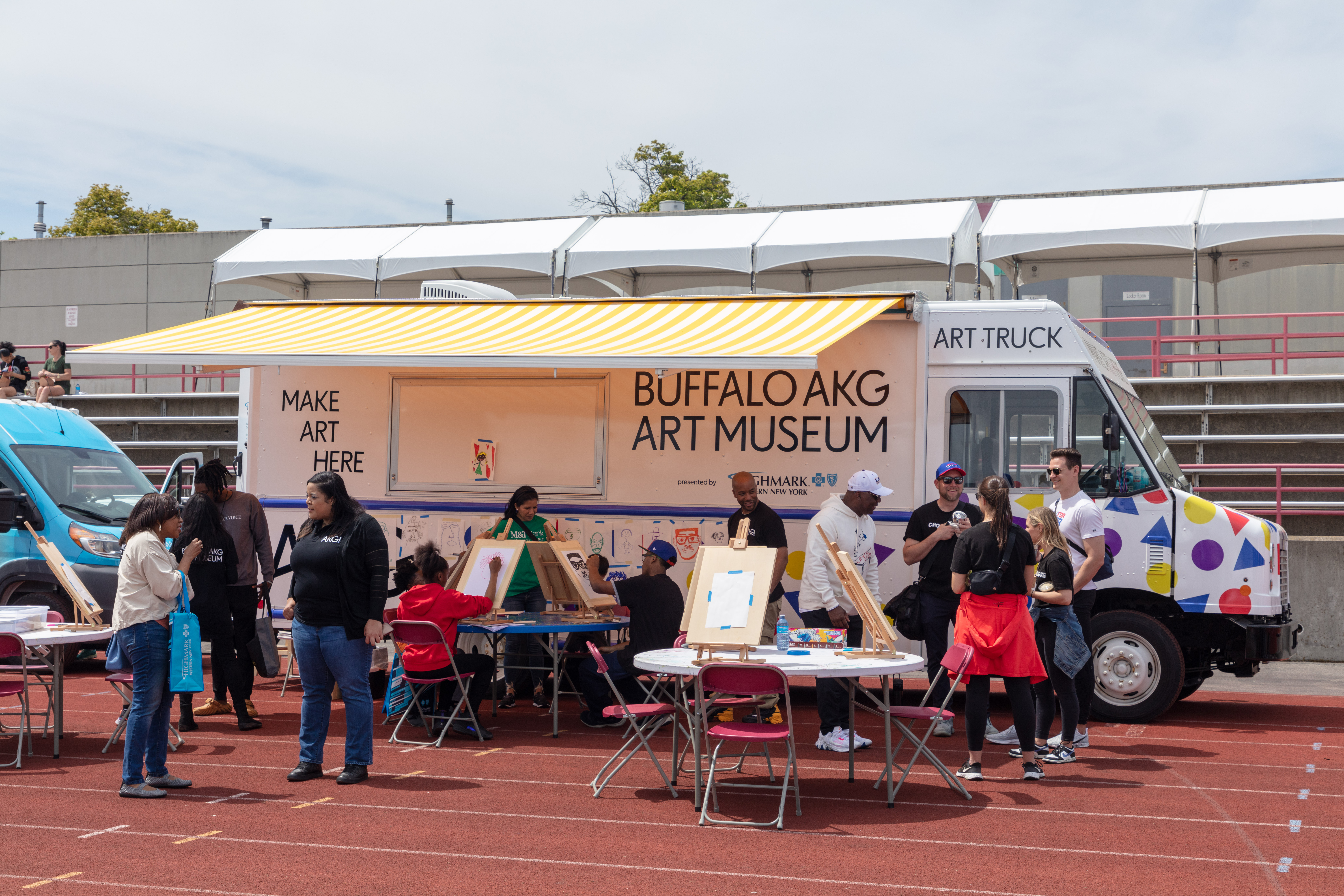A group of people participate in artwork activities at several tables in front of the Buffalo AKG Art truck (that is white with a yellow striped awning) 
