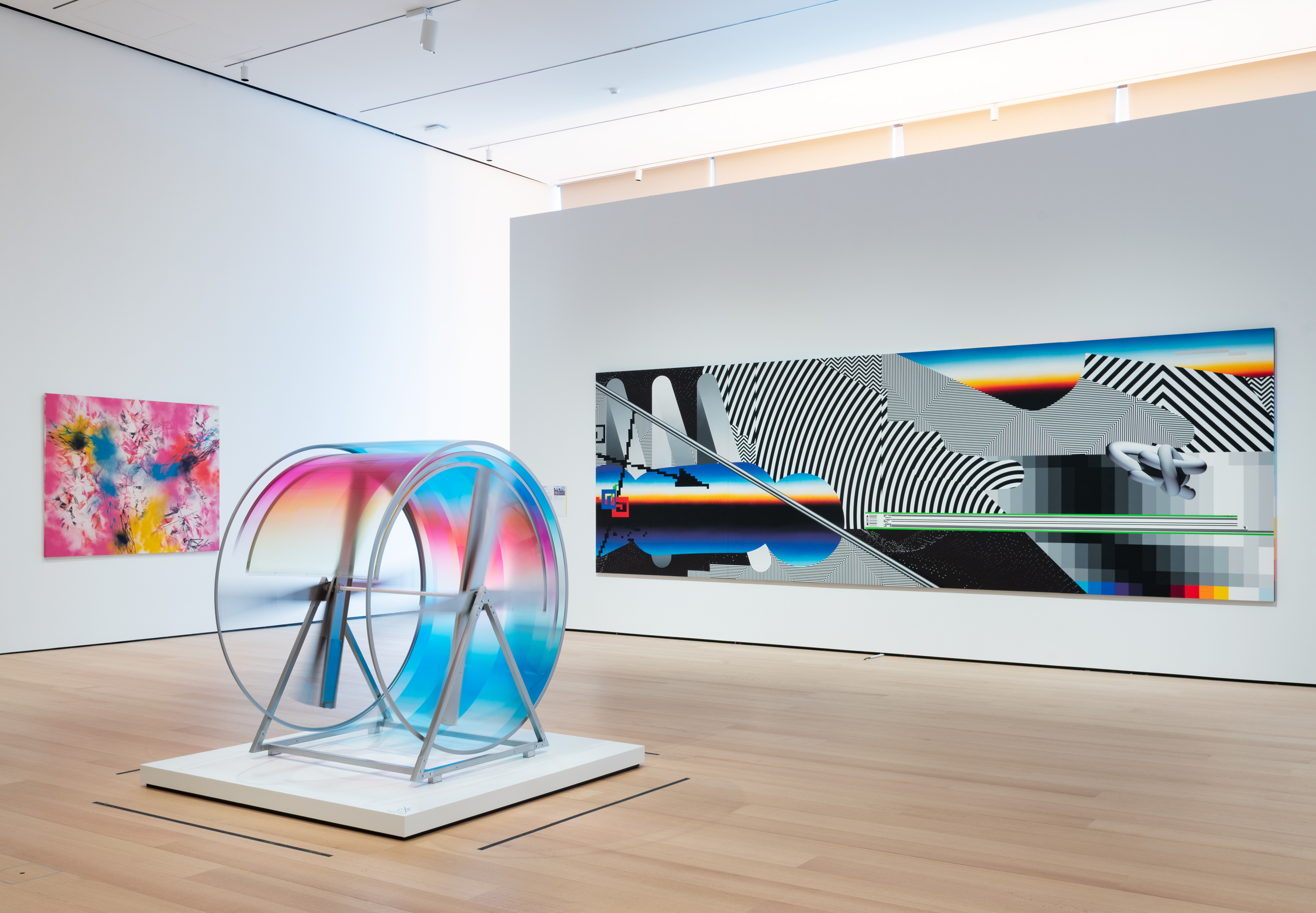 Installation view of a gallery with vibrant paintings and spinning sculpture in the center of the room