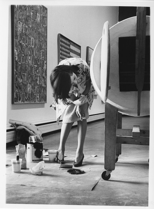 Black and white image of a woman touching up the paint on a wooden sculpture in a gallery