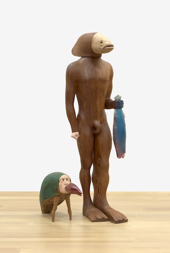 A wood sculpture of a man with a fish head holding a fish in his left arm, his other hand in a fist by his side, at his feet a creature in a hard green shell with a human face and a crab claw extending from its mouth