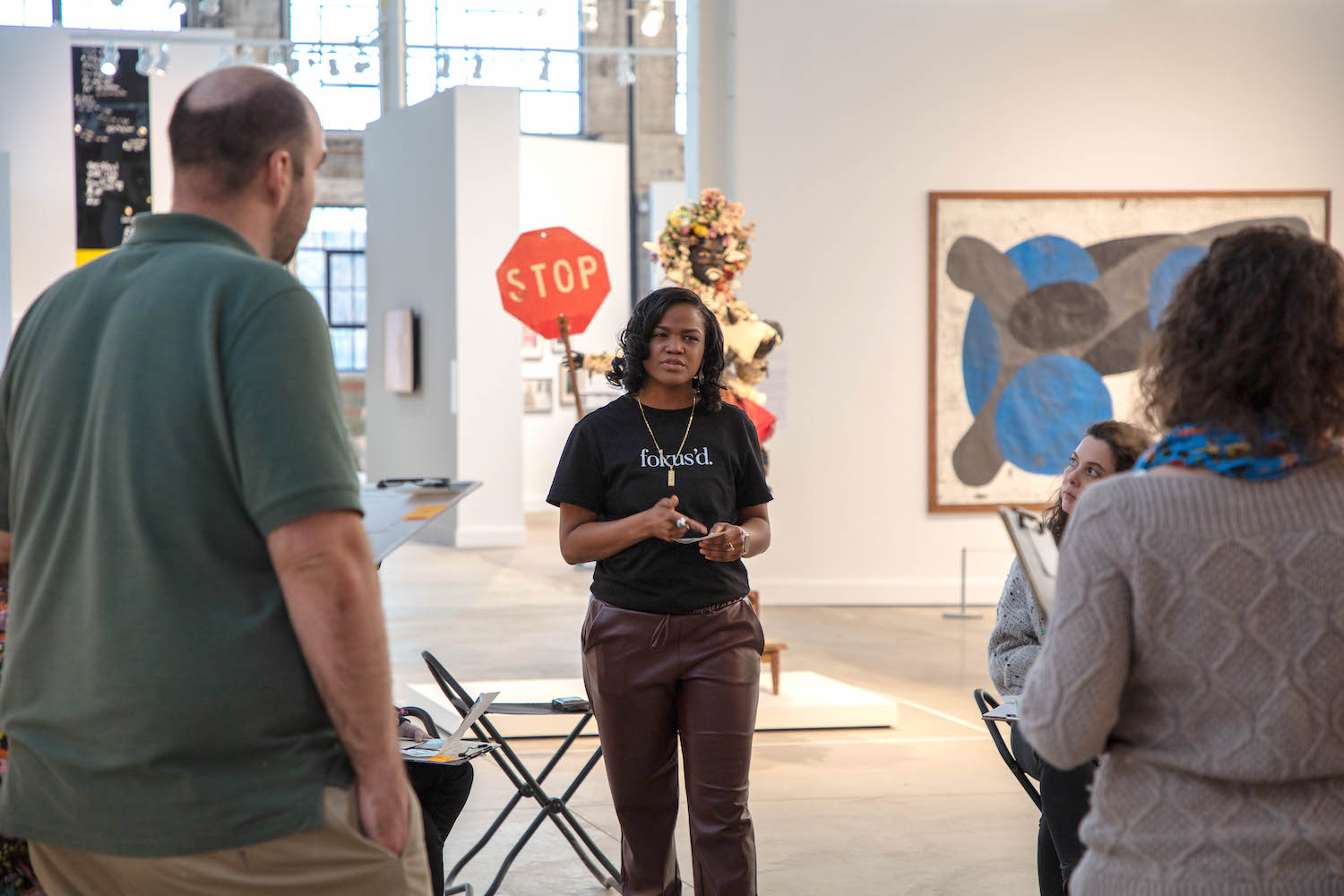 A woman of dark skin tone stands in front of a group speaking and gesturing emphatically