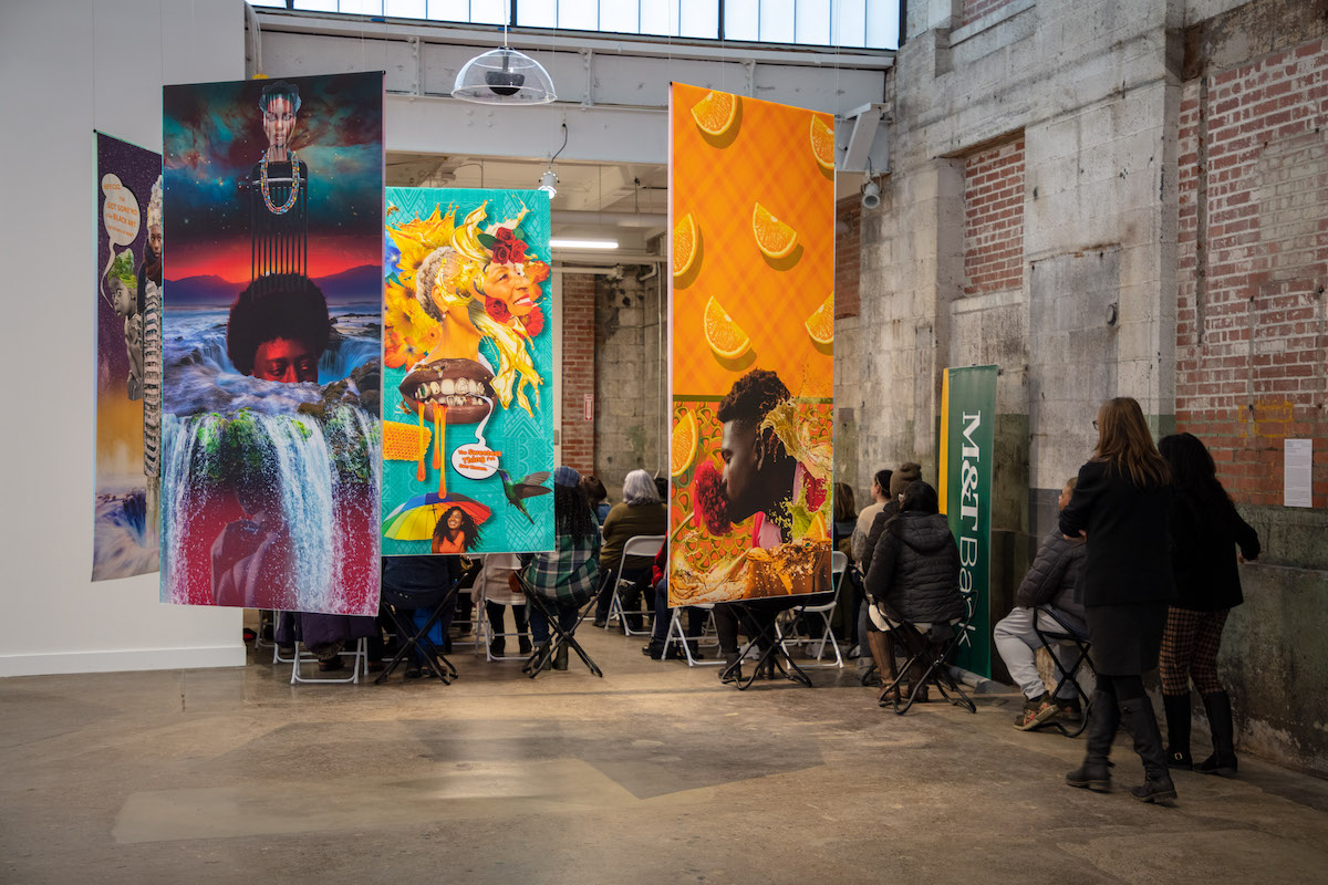 Behind brightly-colored hanging collages people gather in an alcove watching speakers