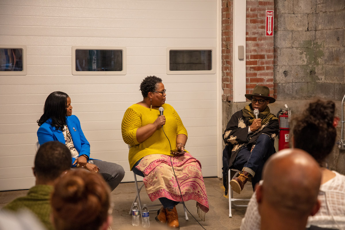 A man of dark skin tone (right) speaks into a microphone with two other speakers