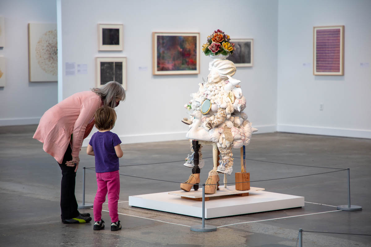 A woman of light skin tone with gray hair bends over to eye level with a young person in pink pants, both looking at sculpture before them