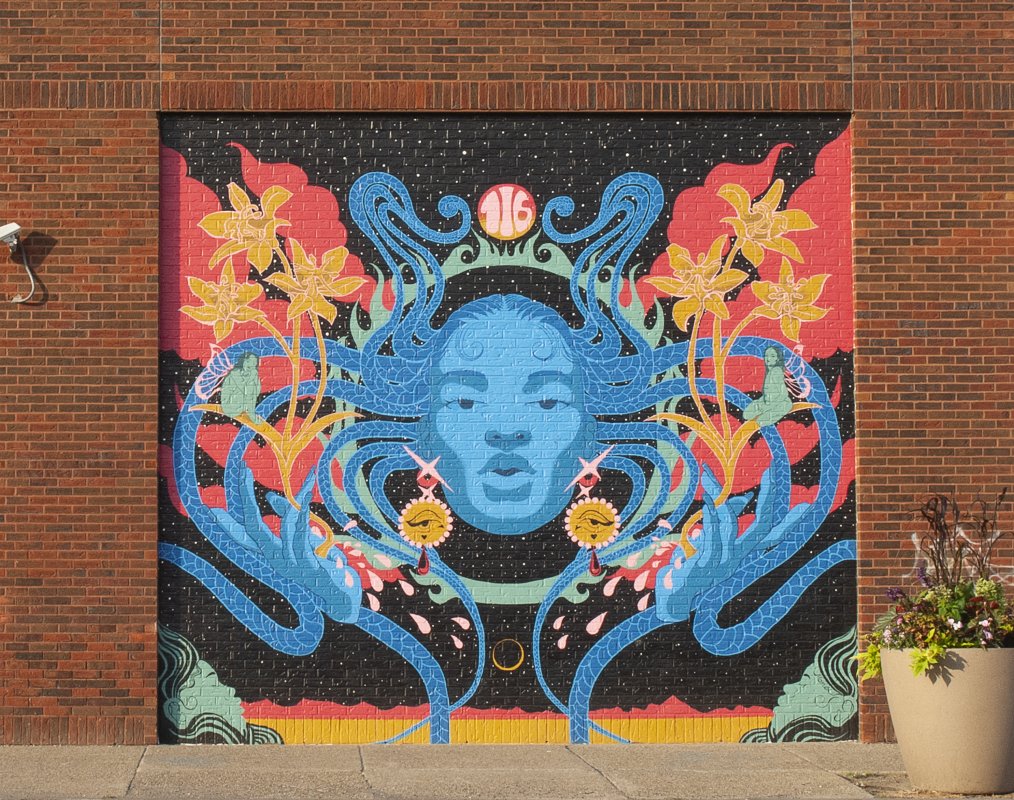 A mural on a brick wall of a woman with blue skin and long blue braids surrounded by florals 