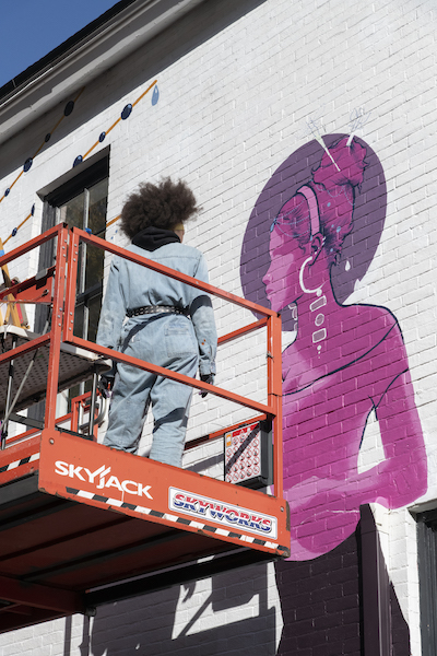 A woman standing on a orange forklift looking at a mural of a woman in progress