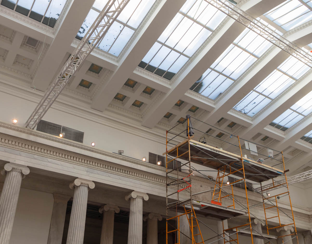 A scaffold rises to a white plaster ceiling in a columned room with a window up above
