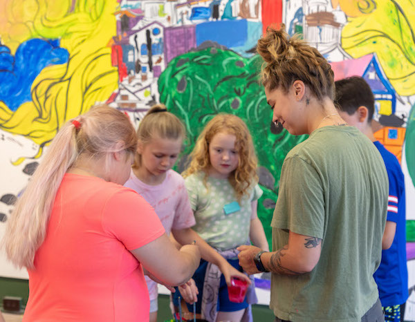 A woman with a green shirt and hair in a bun facing a group of students in front of a colorful mural