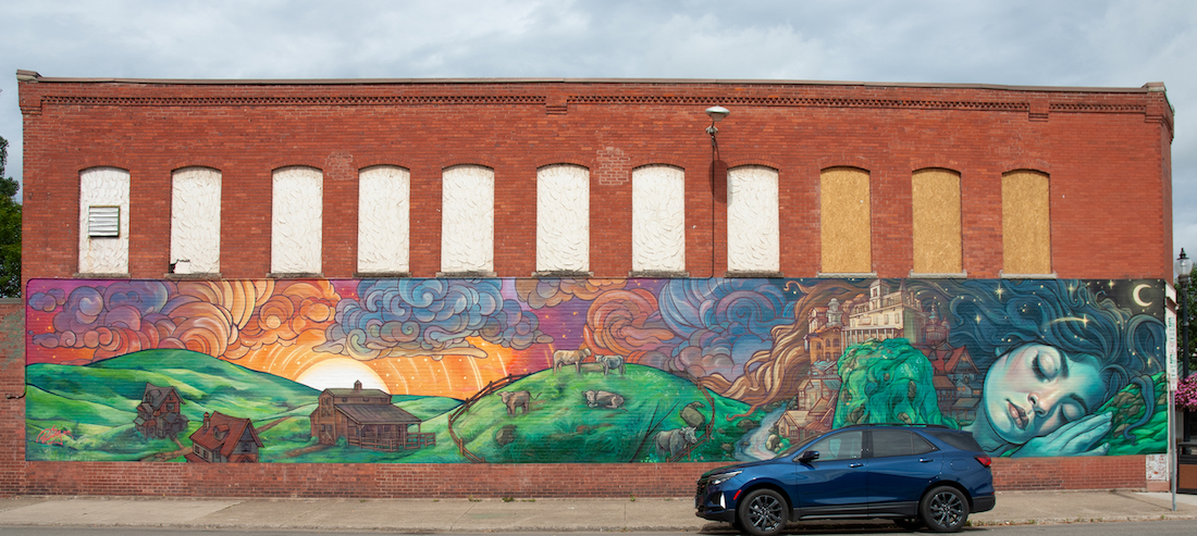 A mural on a brick wall of a green giantess sleeping with a colorful town growing atop her, with a blue SUV parked in front 