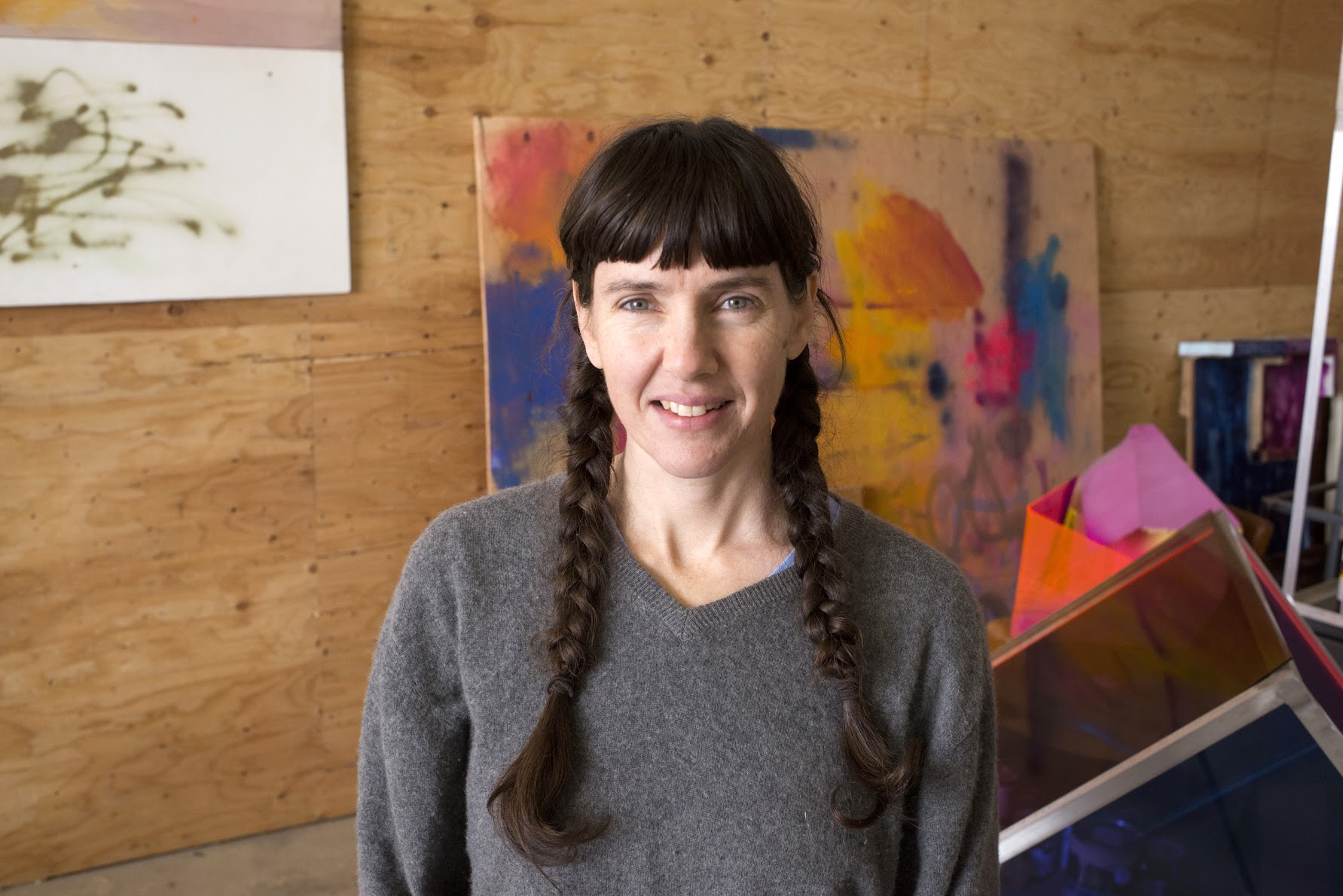 A woman of light skin tone and brown hair in two braids sits facing front, behind her an unvarnished wooden wall against which are leaned a number of paintings