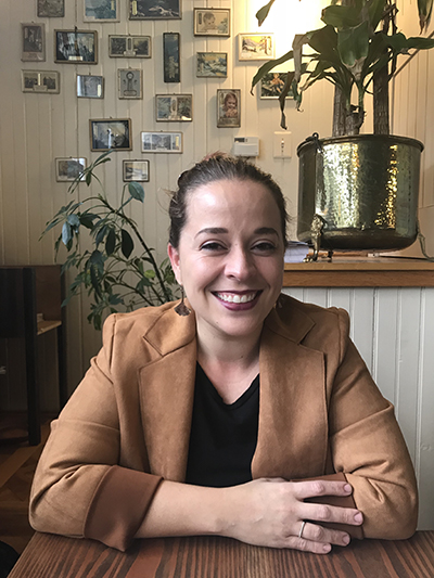 A Latinx woman with her hair pulled back seated at a table with her arms crossed, resting on the table in front of her