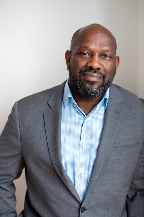 Headshot of a man with dark skin tone wearing a grey suit jacket and blue button up shirt