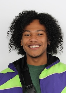 Headshot of a man with long curly dark hair, medium skin tone, smiling and wearing a bright purple and green striped jacket