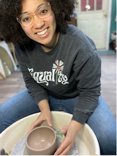 A woman with curly dark hair, medium skin tone, and clear framed glasses, smiling while sitting at a pottery wheel