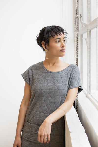 Headshot of a woman with short dark hair, medium skin tone, wearing a grey shirt, leaning against a railing looking out a window