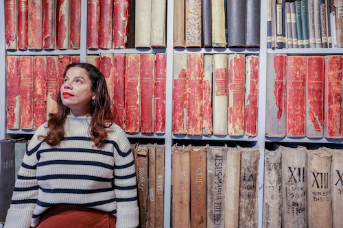 A person with medium-light skin tone and long brown hair looks up and to the right, behind her a row of large, old red books