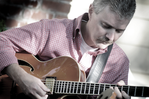 A man with fair skin, a grey mustache and hair, wearing a pink plaid shirt while playing the guitar