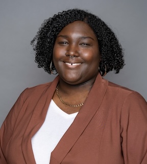 Headshot of a woman with short, dark curly hair, dark skin tone, wearing a brown suit jacket, smiling.