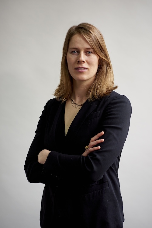A person with shoulder-length blonde hair, folding their arms and looking stoic.