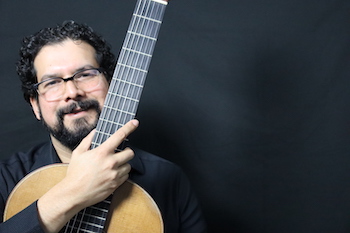 A man with dark hair and fair skin, a dark beard, and glasses holding a guitar close to his face