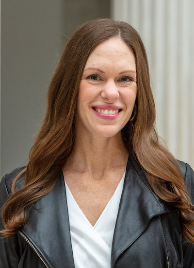 A woman of light skin tone and long, wavy brown hair in a white blouse and leather jacket