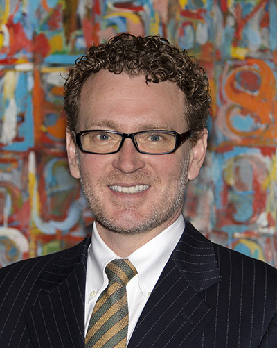 Headshot of a white man with glasses and curly hair in front of a colorful painting