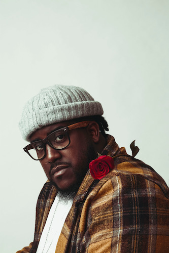 A black man with a grey beanie, glasses, a yellow flannel with a rose on his shoulder looking towards to camera