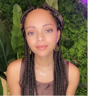 Headshot of a woman with long braided hair, medium skin tone, wearing a brown shirt, smiling in front of a green/plant-filled backdrop