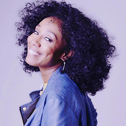A Black woman with long curly hair smiling and wearing a gray-blue jacket