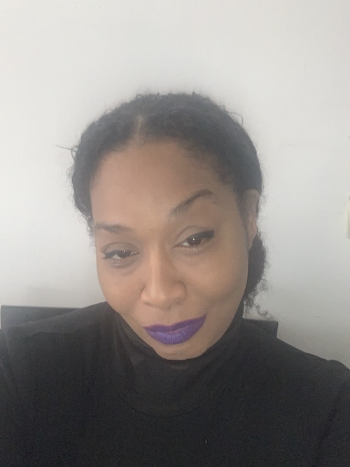 Close up photo of a woman with her dark hair in a bun, medium to dark skin tone, wearing purple lipstick and a black turtleneck shirt