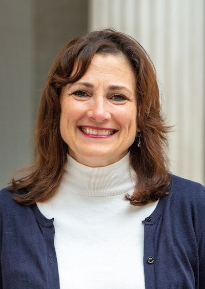 A woman of light skin tone and brown hair in a white turtleneck and blue cardigan