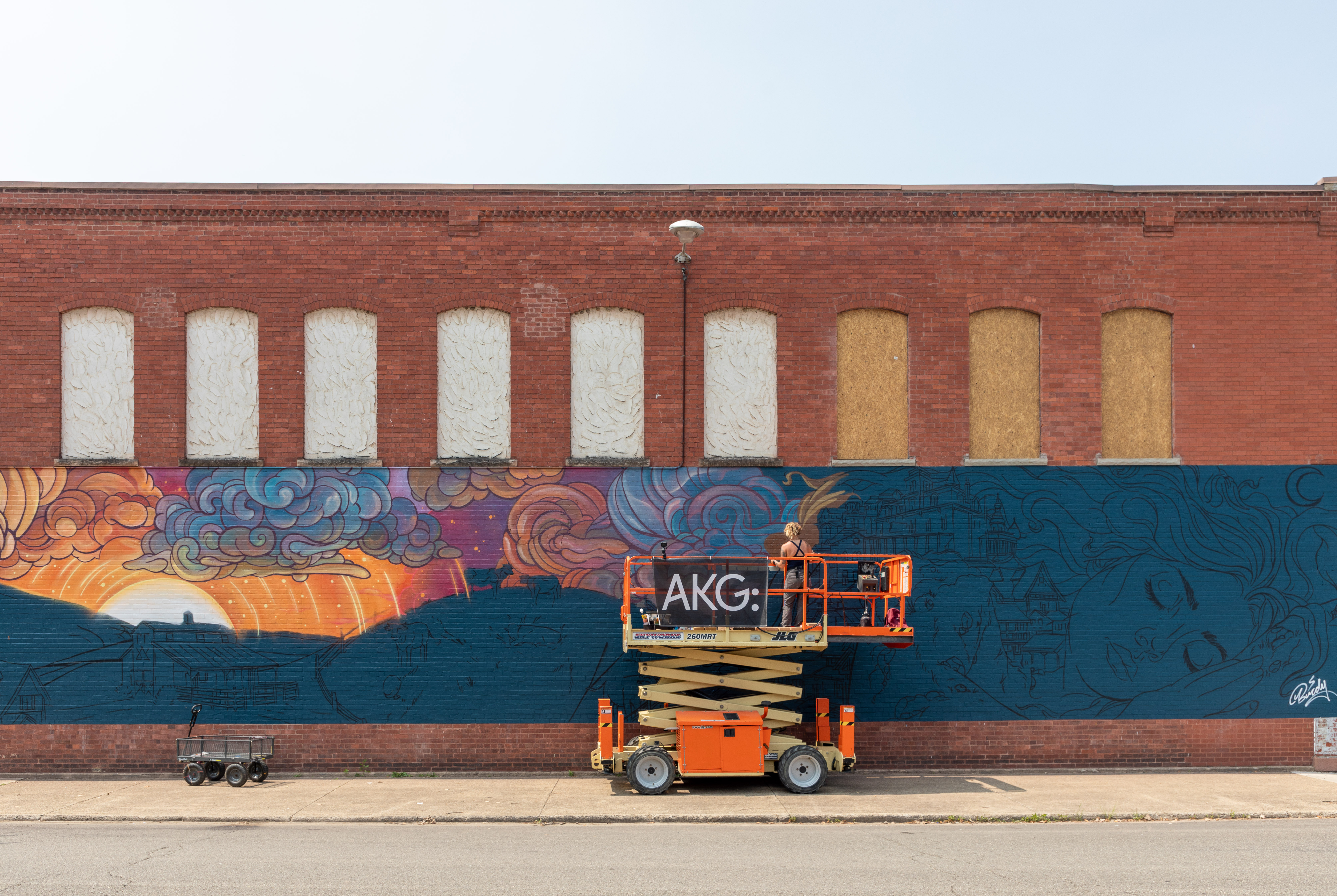 A woman of light skin tone stands on an orange hydraulic lift painting a mural on the side of a red brick building