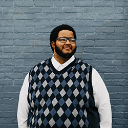 A Black man wearing glasses and an argyle vest over a white long sleeve shirt