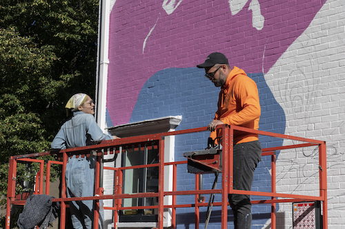 A man in an orange sweatshirt and a woman in a denim jumpsuit standing on a forklift in front of a mural in progress