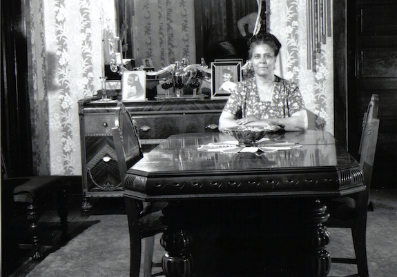 A black and white photo of a woman of medium-dark skin tone sitting at a dining room table, a dining hutch with mirror and assorted photographs on top of it behind her