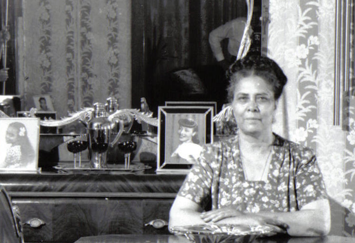 Detail image of black and white photo featuring older woman of medium-dark skin tone seated at table, in mirror above her the cameraman is visible