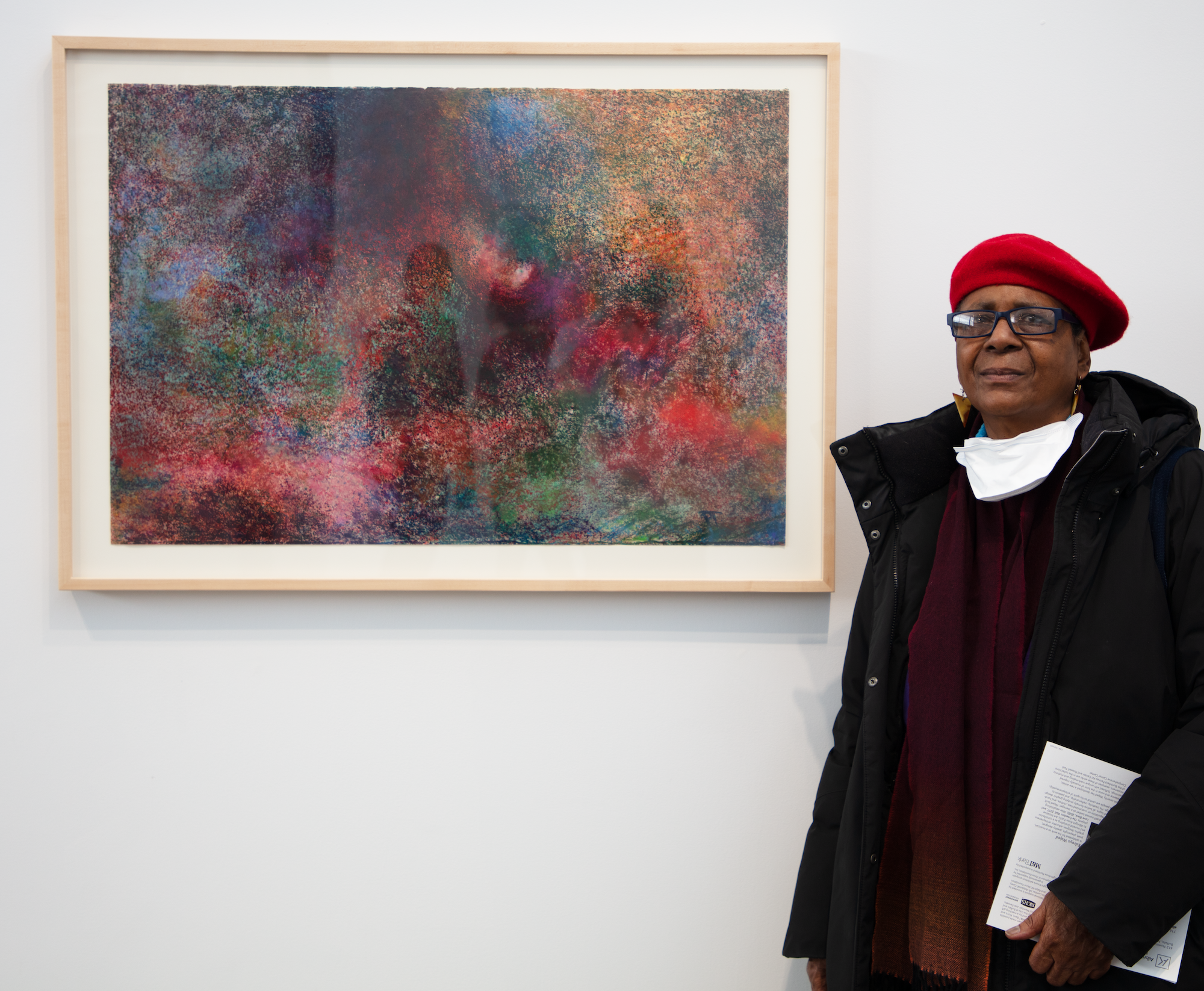 A woman of medium dark skin tone in red beret stands in front of an artwork