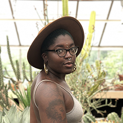 An African American person with black glasses and a brown hat looks at the camera over their shoulder