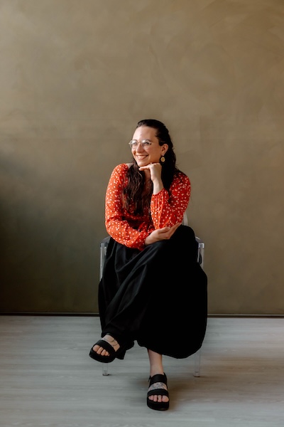 A person seating with their legs crossed and resting their head in their hand, smiling, and looking away from the camera 