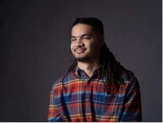 Headshot of a man with long dark braided hair pulled away from his face, medium skin tone, wearing a plaid shirt, looking away from the camera