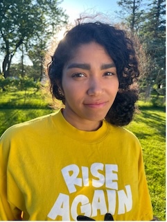 Photo of a woman outdoors with long dark hair pulled into a bun, medium skin tone, wearing a yellow shirt that says "Rise Again" 