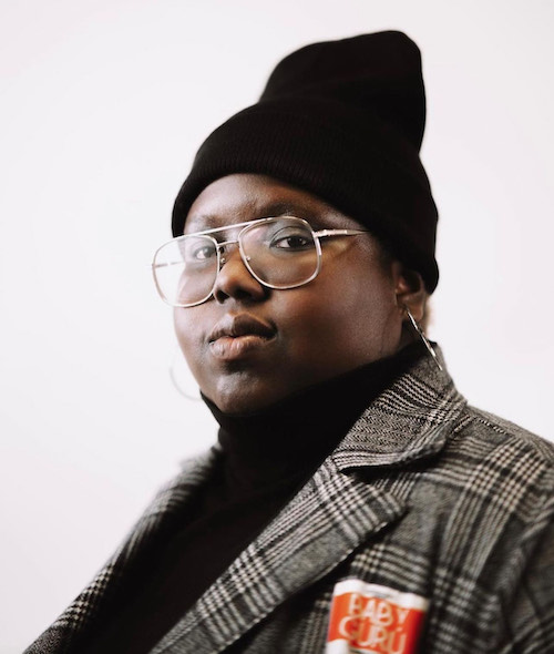 Headshot of a woman with a dark skin tone, large framed glasses, a black beanie, black turtleneck, and plaid dress jacket