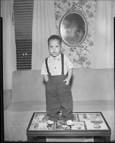 A black and white photo in which a young boy of medium dark skin tone in overalls stands on a coffee table
