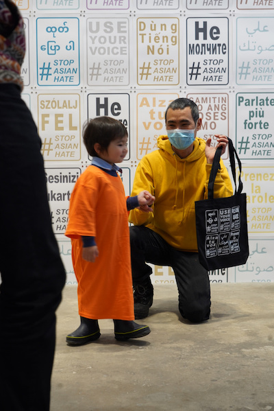 A young boy in a large orange shirt with his father kneeling beside him, both of Asian descent, stand in a gallery space with "Use Your Voice #StopAsianHate" prints lining the walls