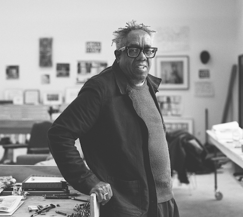Black and white photo of a black man wearing glasses leaning against a desk in an art studio