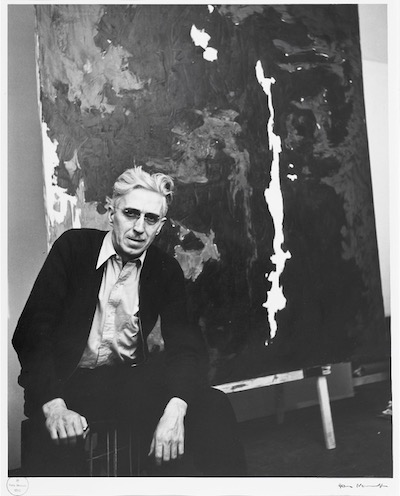 Black and white photograph of man in black jacket sitting with elbows resting on his knees, a painting behind him