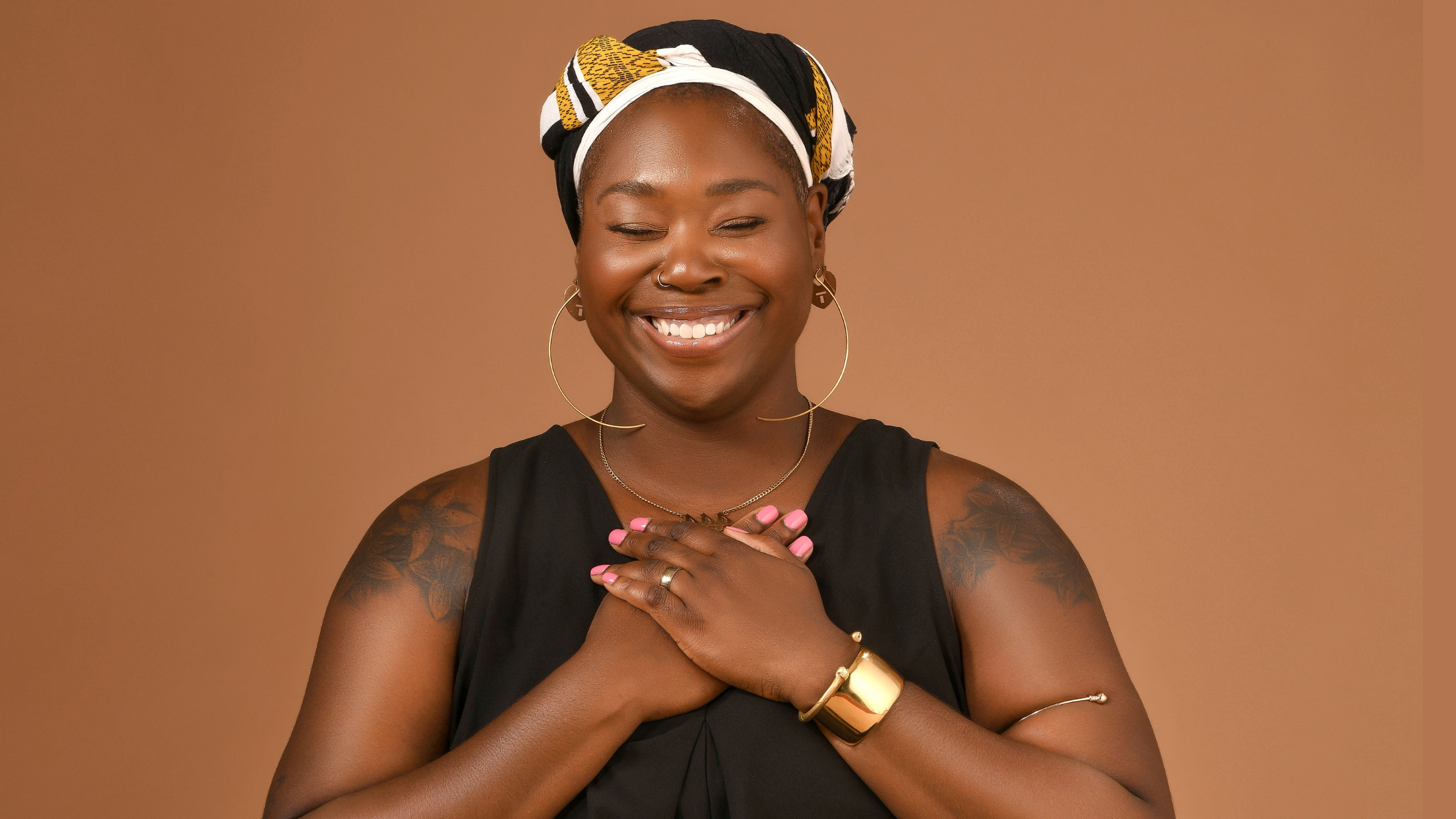 An image of a smiling woman with a colorful headwrap and tattoos on her shoulders