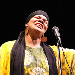 A Black woman singing into a microphone, wearing a black head wrap and large circular earrings