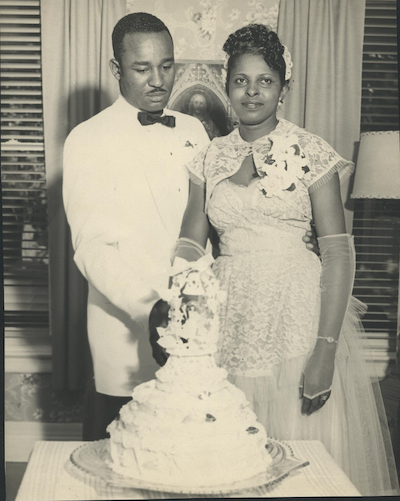 A black and white photo showing a bride an groom of dark skin tone in suit and wedding gown cutting a wedding cake